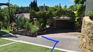 Une allée menant à une résidence, avec un mur de pierre sur la droite et un jardin agrémenté de diverses plantes et d’une statue.