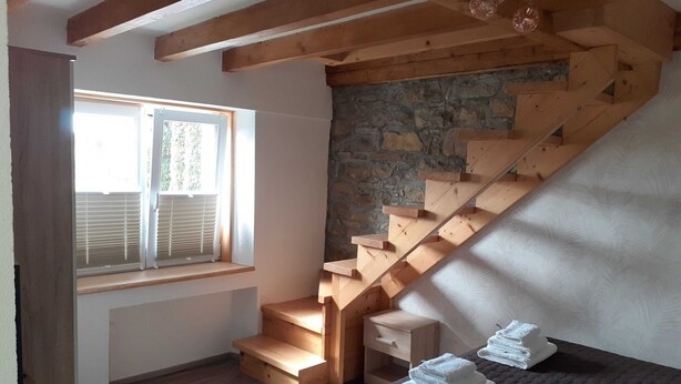 Une chambre avec un escalier en bois, une fenêtre avec des stores, un lit recouvert d  ’une couverture sombre et un mur en pierre.