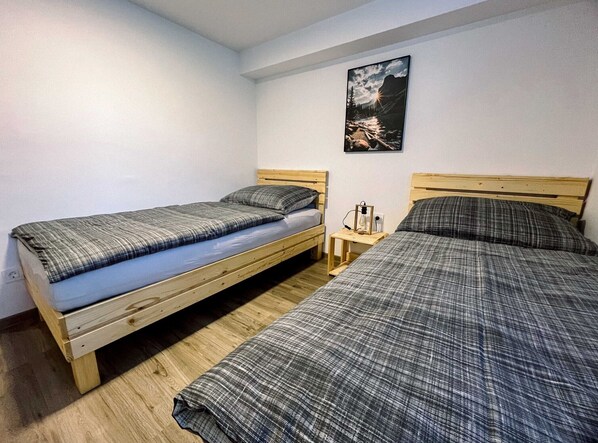 A room with two wooden beds, one with a blue mattress and the other with a grey mattress. There is a small wooden nightstand with a lamp between the beds.