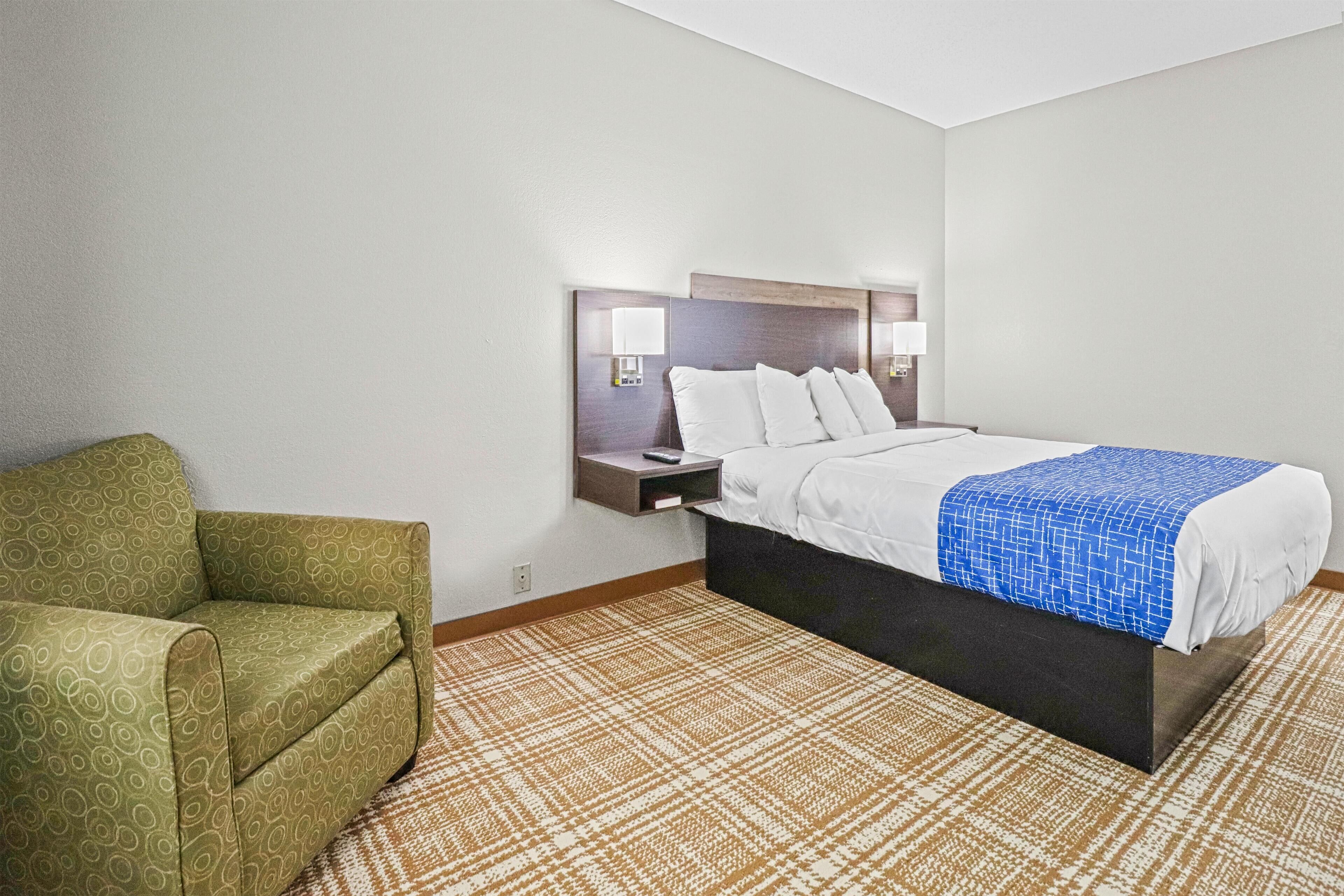 Una habitación de hotel con una cama, una mesita de noche, un sillón verde y una alfombra estampada.