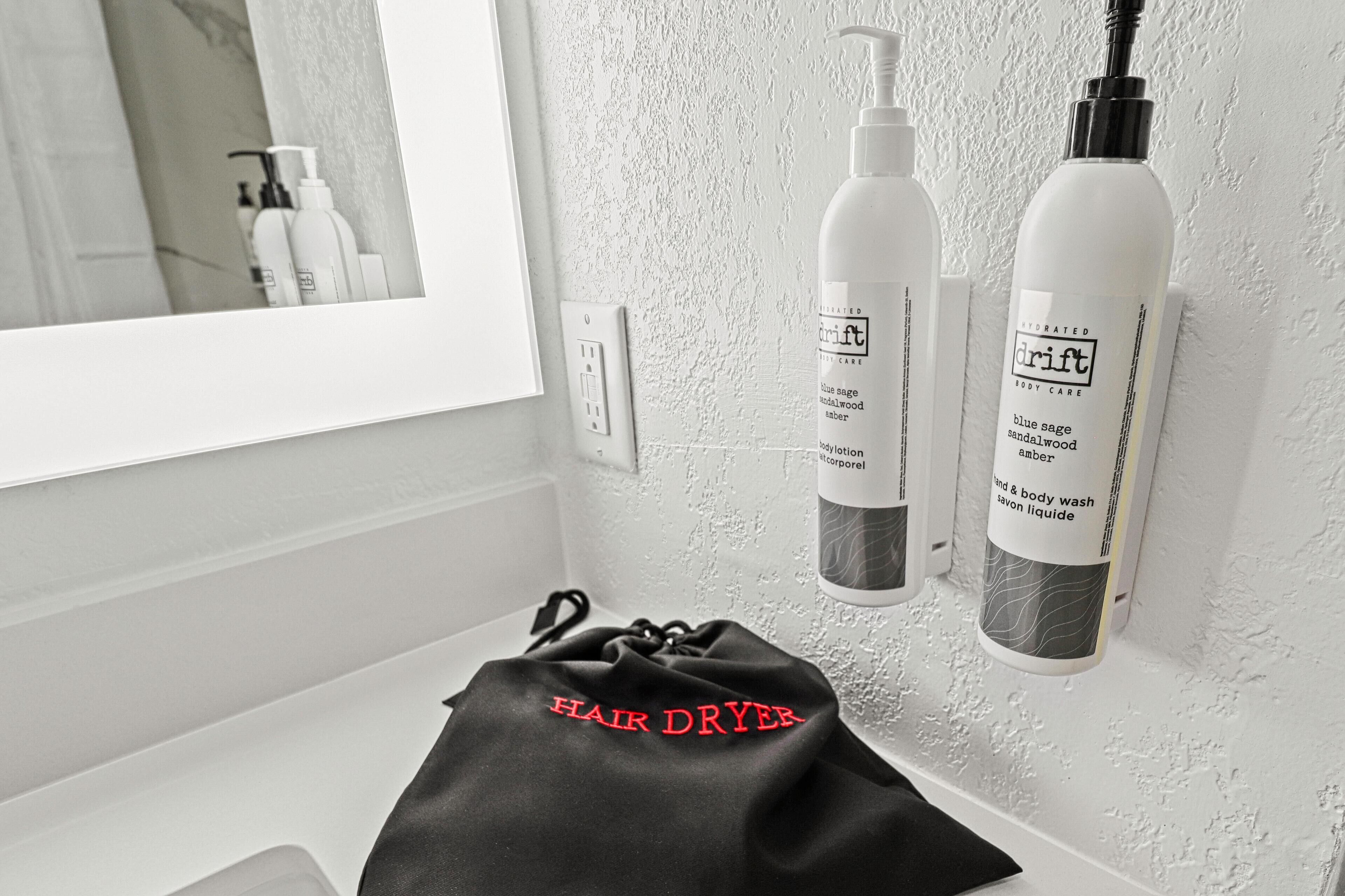 Een badkamer met een witte wastafel, een zwarte handdoek met de tekst 'HAIR DRYER' en twee flesjes 'drift' verzorgingsproducten voor het lichaam.