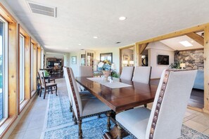 A spacious dining area with a long wooden table and upholstered chairs, a kitchen in the background, and a stone wall.