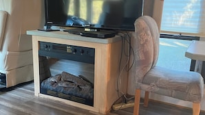 Een compact RV-interieur met een flatscreen televisie op een houten entertainmentmeubel, een comfortabele zithoek en een raam met uitzicht op de natuur.