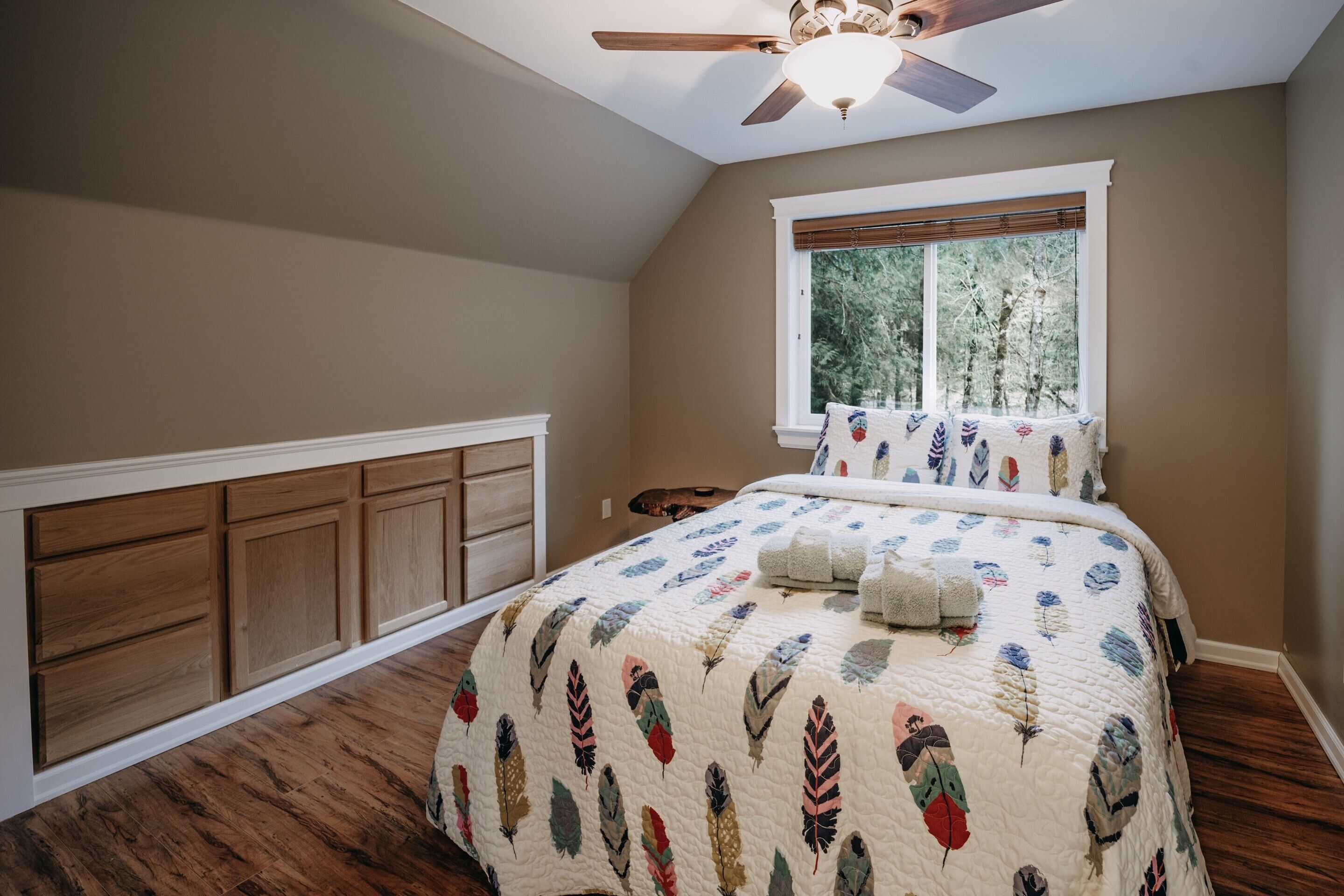A bedroom with a wooden floor, a bed with a feather-patterned quilt, a ceiling fan, and a window with a view of trees.
