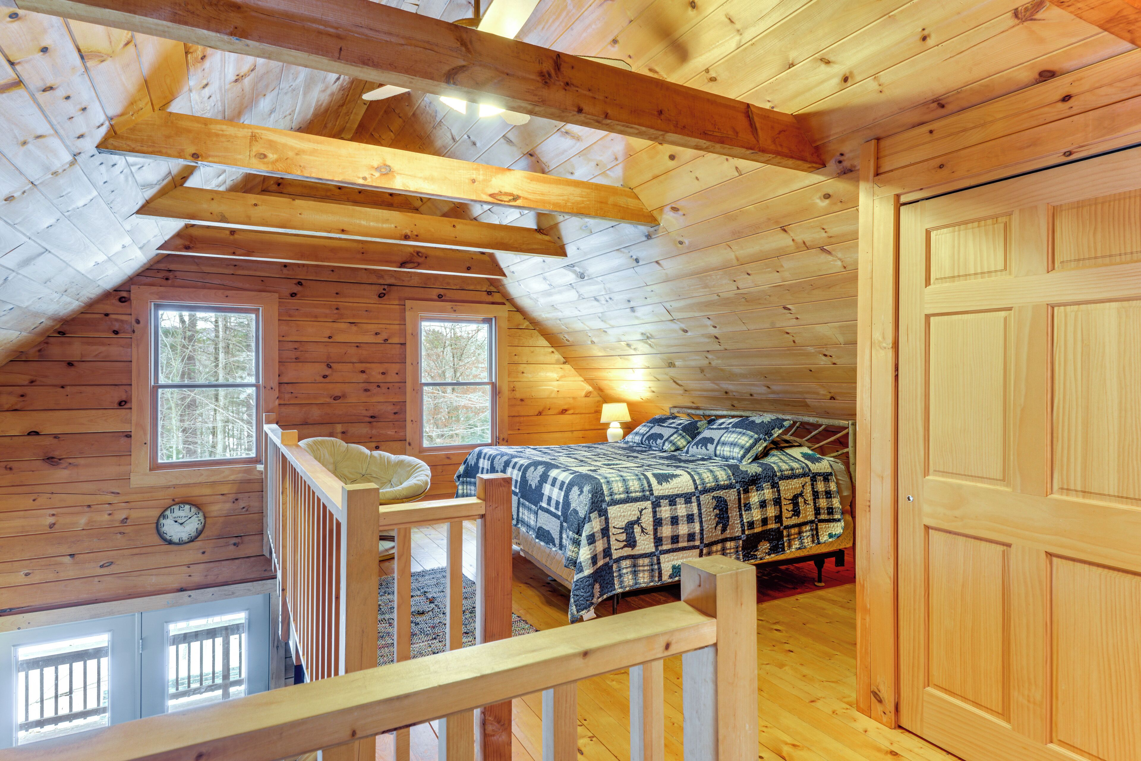 A cozy attic bedroom with a wooden ceiling, a bed with a checkered blanket, a small seating area, and a window with a view of trees.