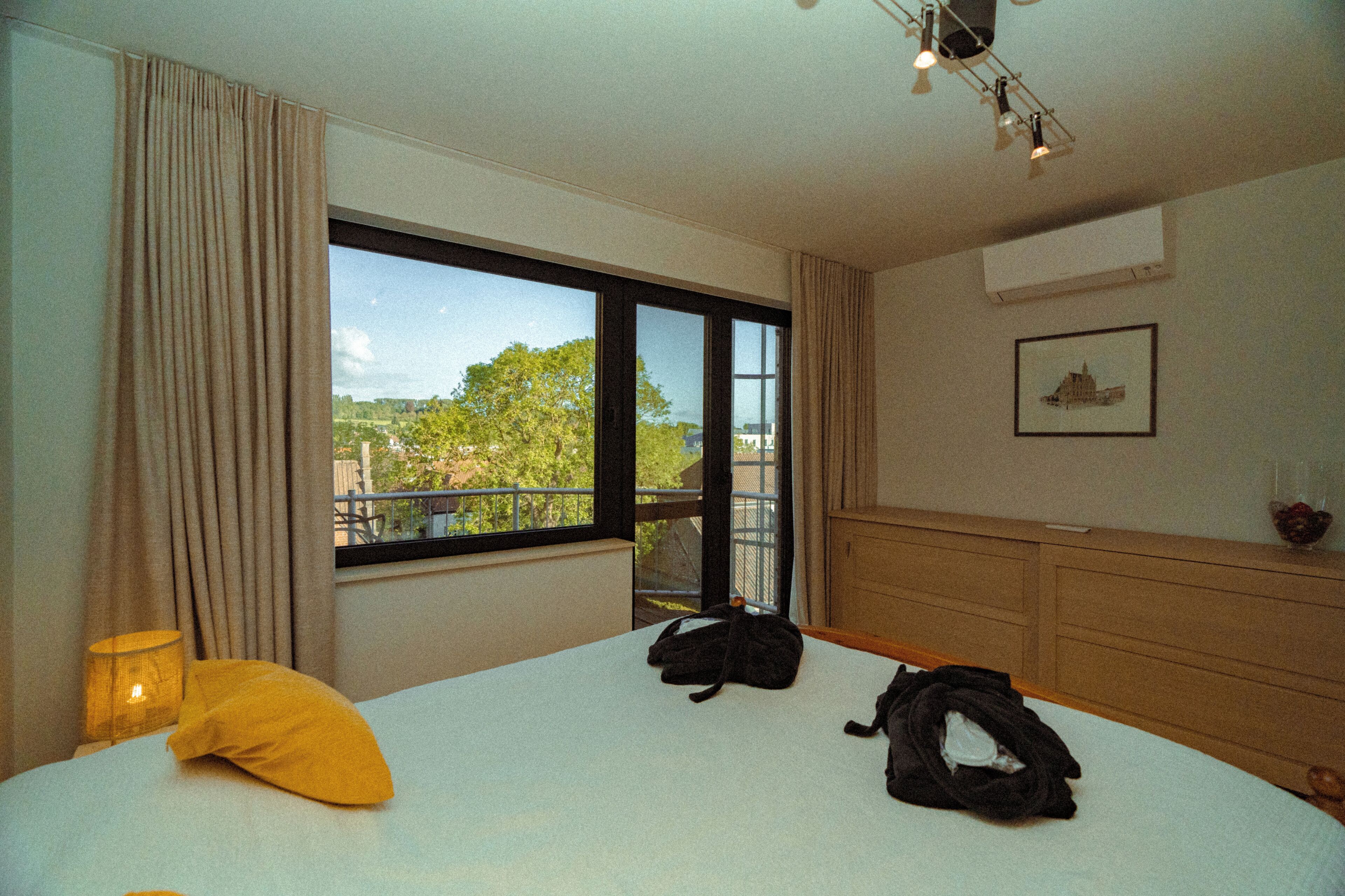 A hotel room with a large window, a bed, a bedside lamp, and a wooden dresser.