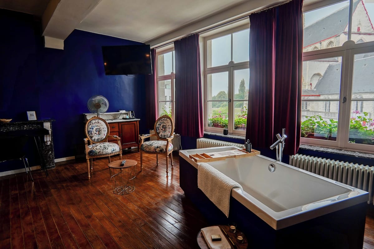 Une salle de bain avec une grande baignoire, deux chaises, un ventilateur et une fenêtre avec des rideaux.