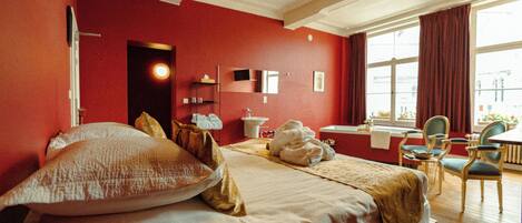 A hotel room with a large bed, a desk, and a chair. There is a window with curtains, a mirror, and a sink.