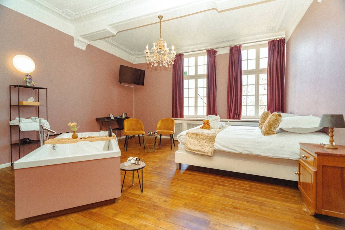 Une chambre d’hôtel avec un grand lit, un bureau, une chaise, une coiffeuse avec un miroir et un lustre.