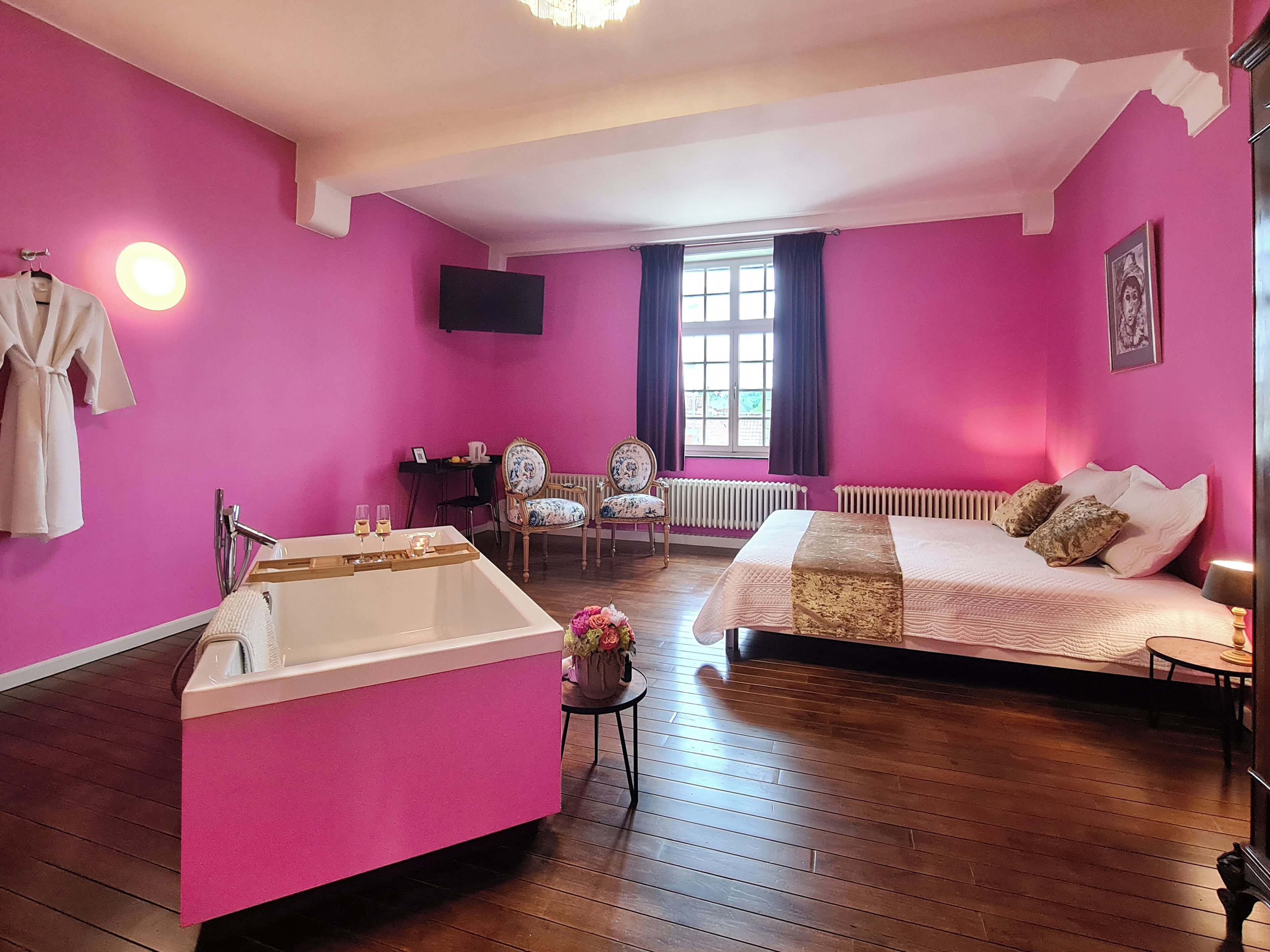 A room with a pink wall, a bathtub, a bed, a chair, a table, and a window.