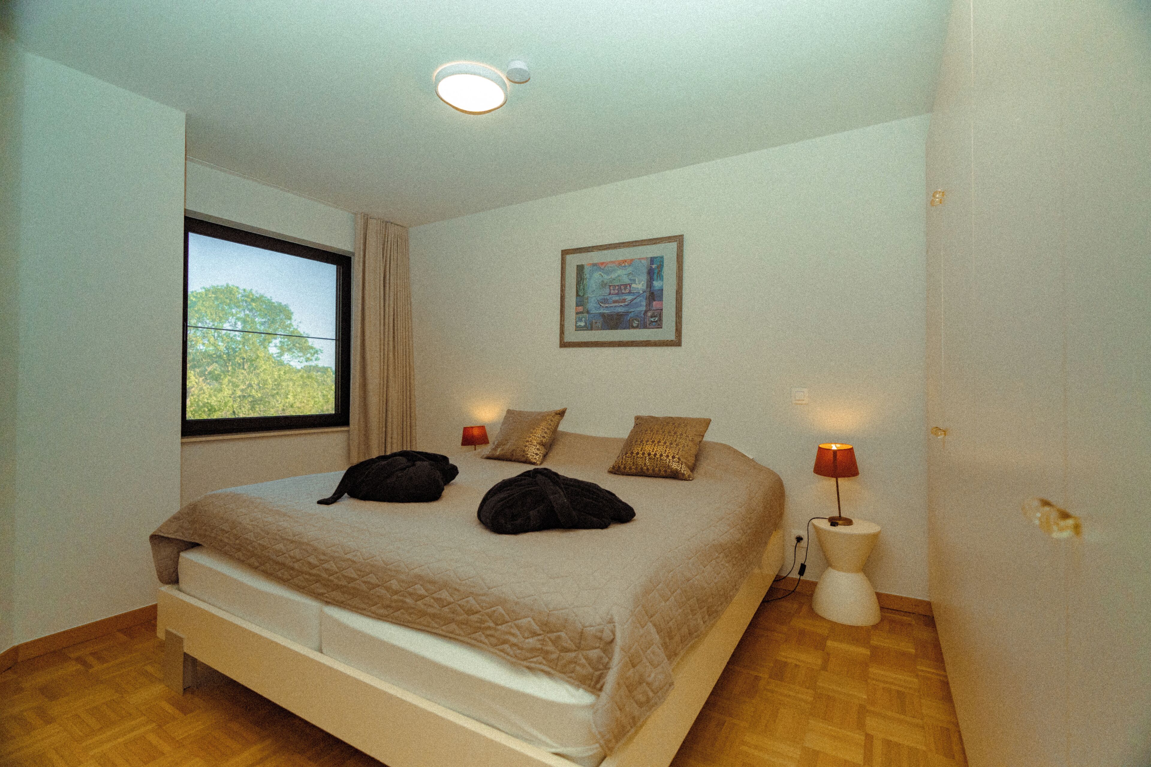 A bedroom with a large bed, two bedside lamps, a window with curtains, and a painting on the wall.
