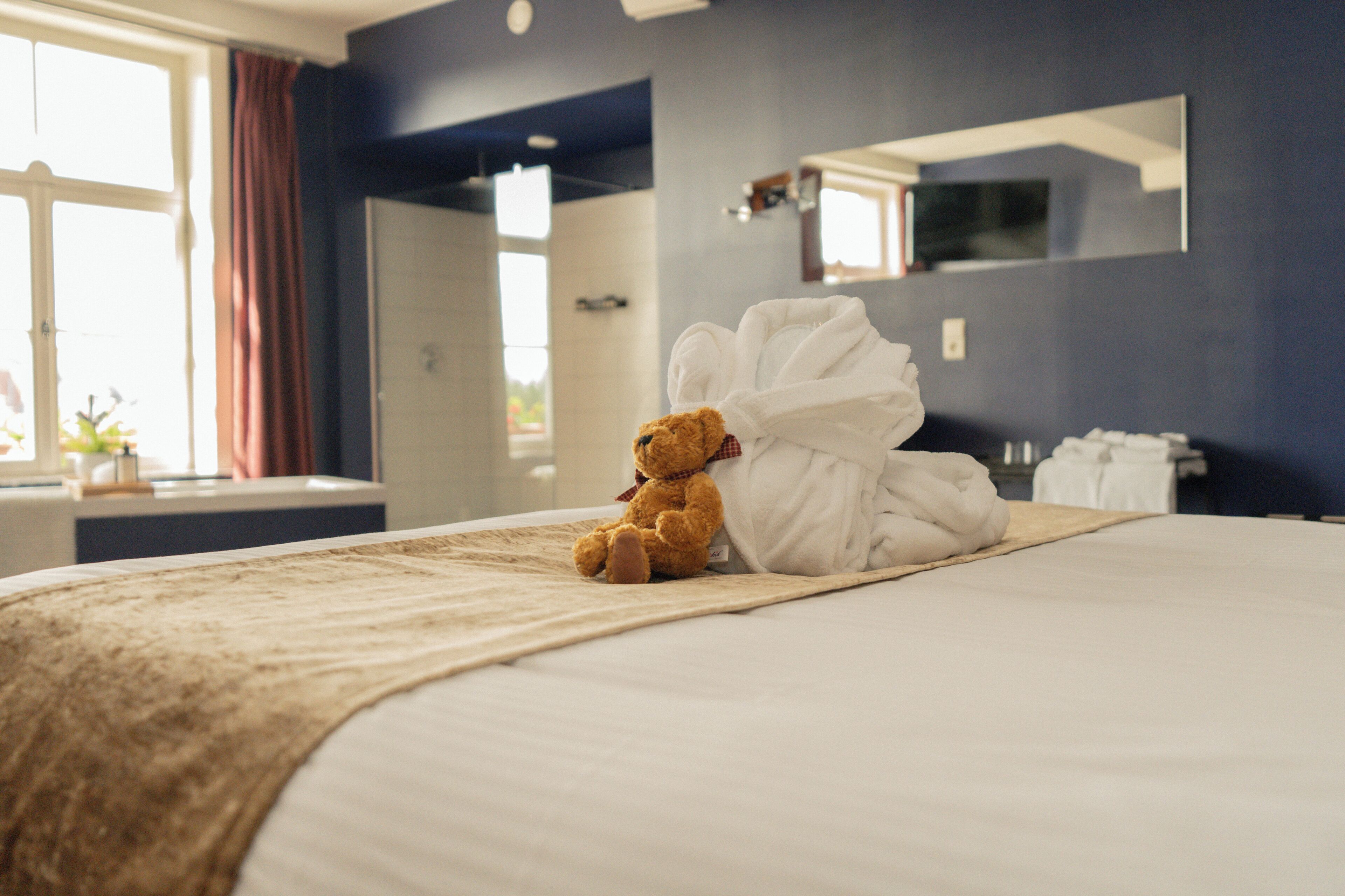 A neatly made bed with a teddy bear and a white bathrobe on it, a window with curtains, and a mirror on the wall.