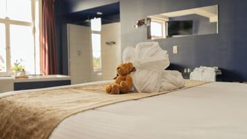 A neatly made bed with a teddy bear and a white bathrobe on it, a window with curtains, and a mirror on the wall.