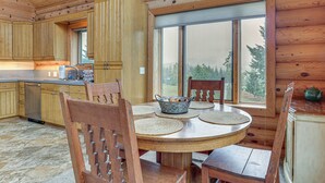 A wooden interior with a round dining table, chairs, a kitchen area, and a large window.