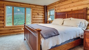 A wooden log cabin bedroom with a large bed, a nightstand, and a window with a view of a forest.