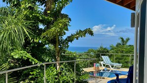 Un balcon avec vue sur l’océan, entouré d’une végétation luxuriante et donnant sur un espace piscine agrémenté de chaises longues.