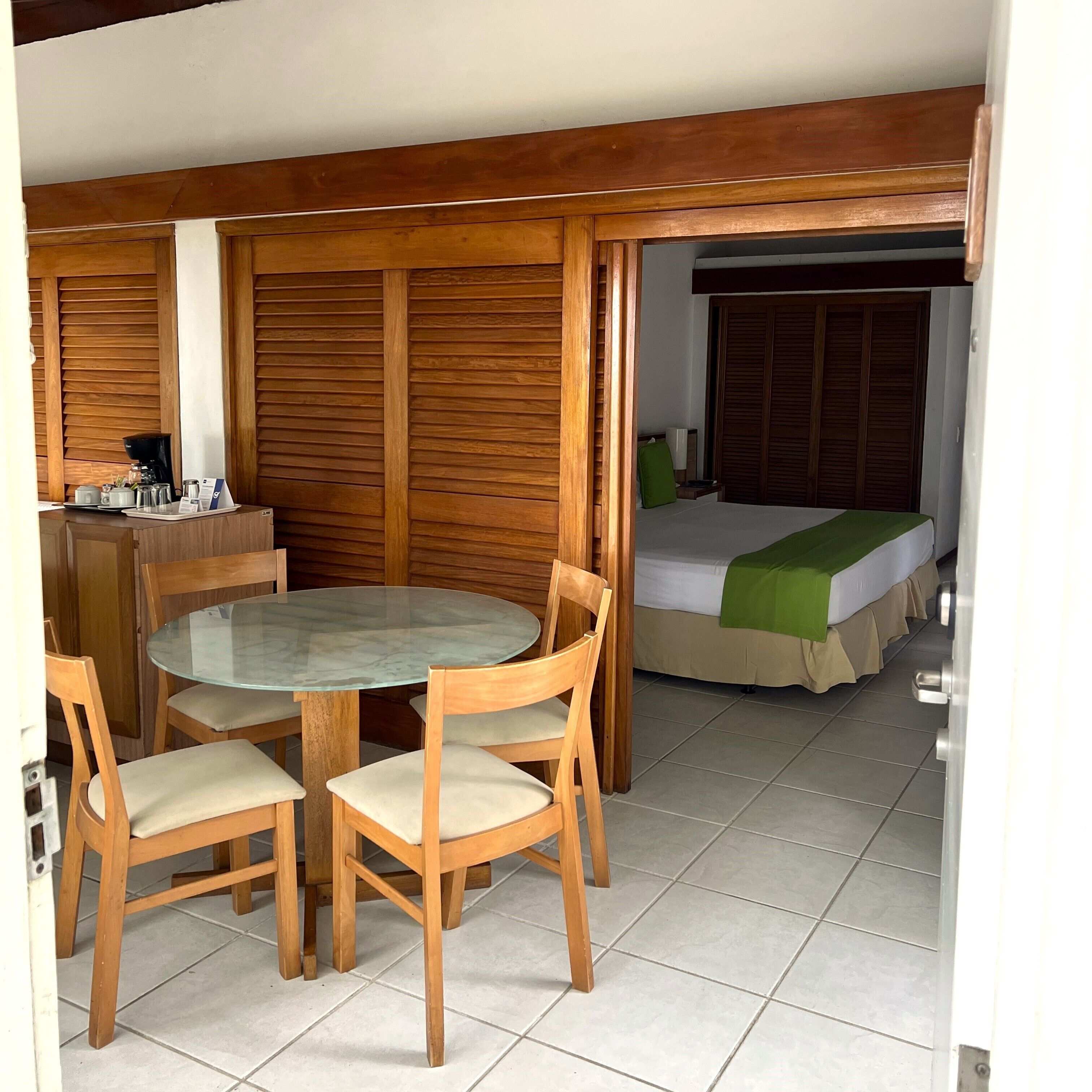 Una habitación con una mesa redonda de vidrio, sillas de madera, una cama con una manta verde y un armario de madera.