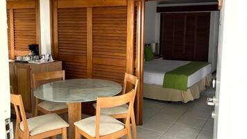 Una habitación con una mesa redonda de vidrio, sillas de madera, una cama con una manta verde y un armario de madera.