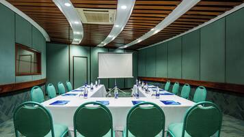 Un salón de conferencias con una larga mesa, sillas color turquesa y una pantalla blanca al frente.