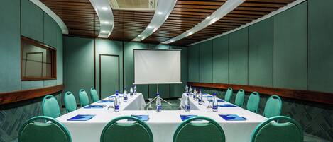 A conference room with a long table, teal chairs, and a white screen at the front.
