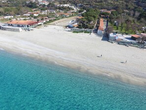 An aerial view of a beach with clear turquoise waters and a sandy shore. There are buildings with red roofs and a few trees in the background.