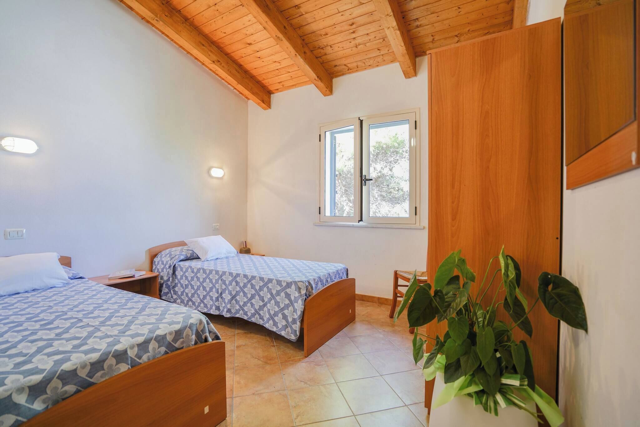 A bedroom with two beds, wooden ceiling beams, a window with a view, and a potted plant.