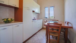 A kitchen with white cabinets, a wooden table, and chairs. There is a window with a view of greenery outside. - Villaggio Residence Emmesse Trilocal 3 Rooms (Zambrone)