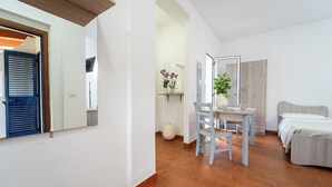 A bedroom with a bed, a desk, and a chair. There is a mirror on the wall and a door with blue shutters. - Appartamenti Standard (Zambrone)