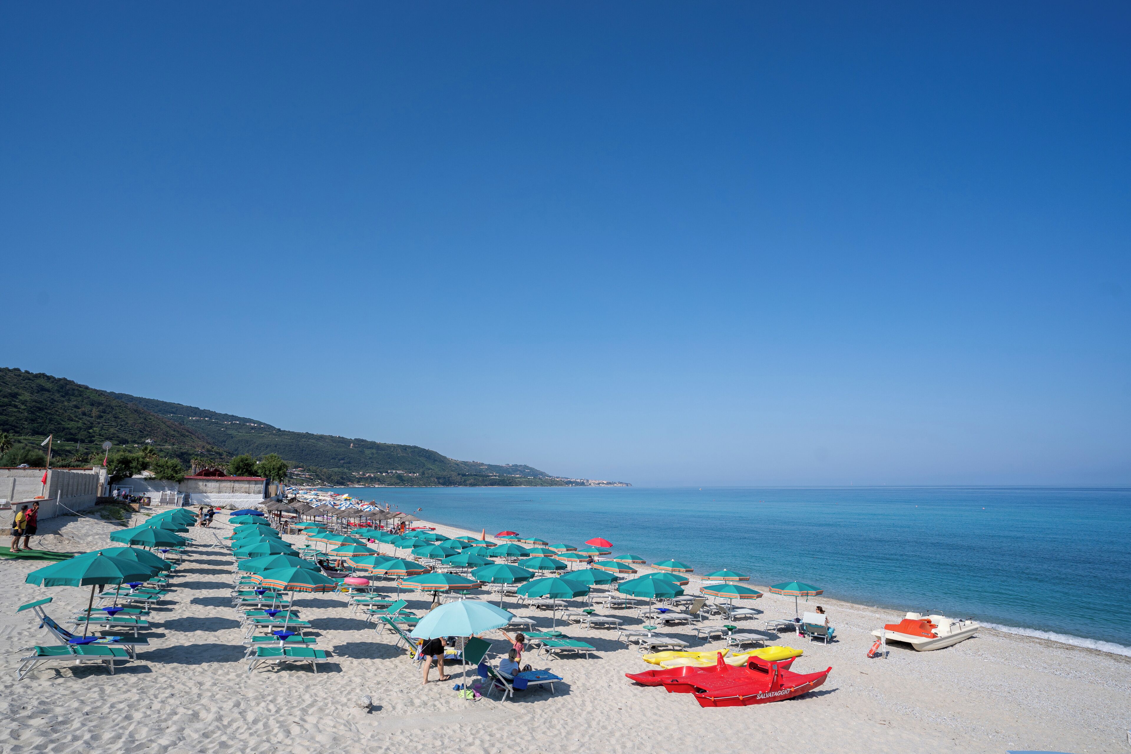 A beach with rows of sun loungers and umbrellas, a boat on the sand, and a clear blue sky.