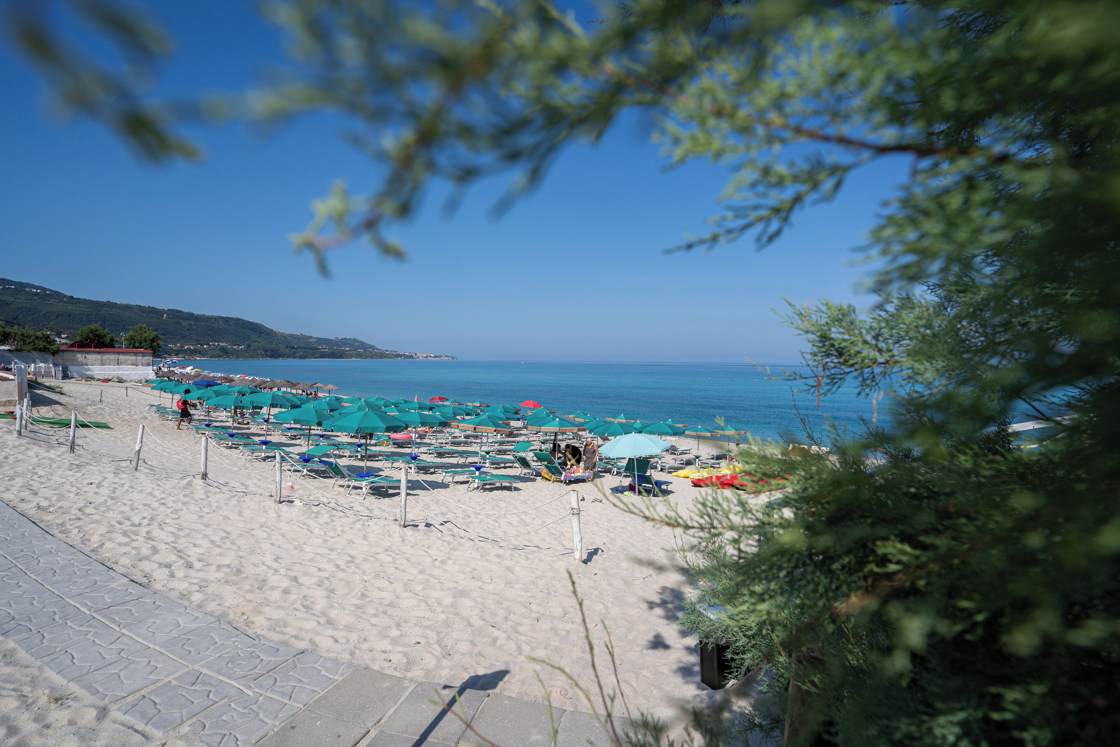 A beach with rows of umbrellas and sun loungers, a clear blue sky, and a hilly landscape in the background.