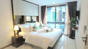 A hotel room with a large bed, a bedside table with a lamp, a window with curtains, and a view of buildings outside. - B307 - 1 Bedroom Condo Pool View Near Ao Nang Beach (Krabi)
