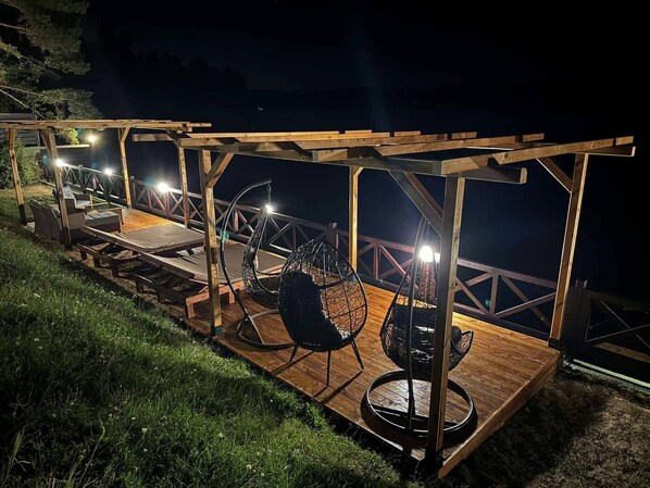 Terraza de madera al aire libre con asientos y sillones colgantes, iluminada por luces colgantes.