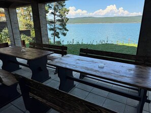 An outdoor seating area with wooden benches and tables, overlooking a lake with a distant shoreline and hills.