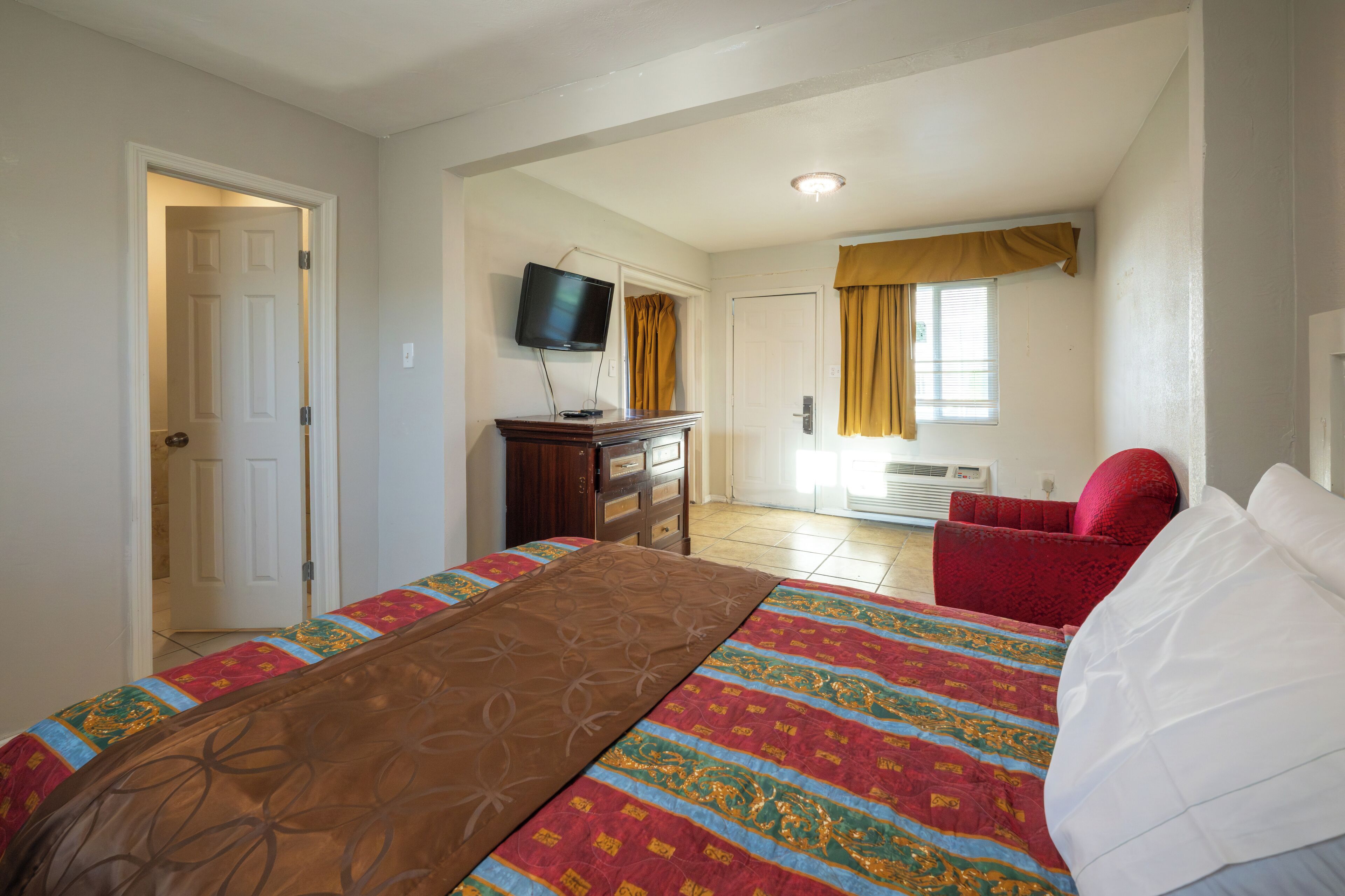 Un dormitorio con una cama, televisor, una silla roja y una ventana con cortinas.
