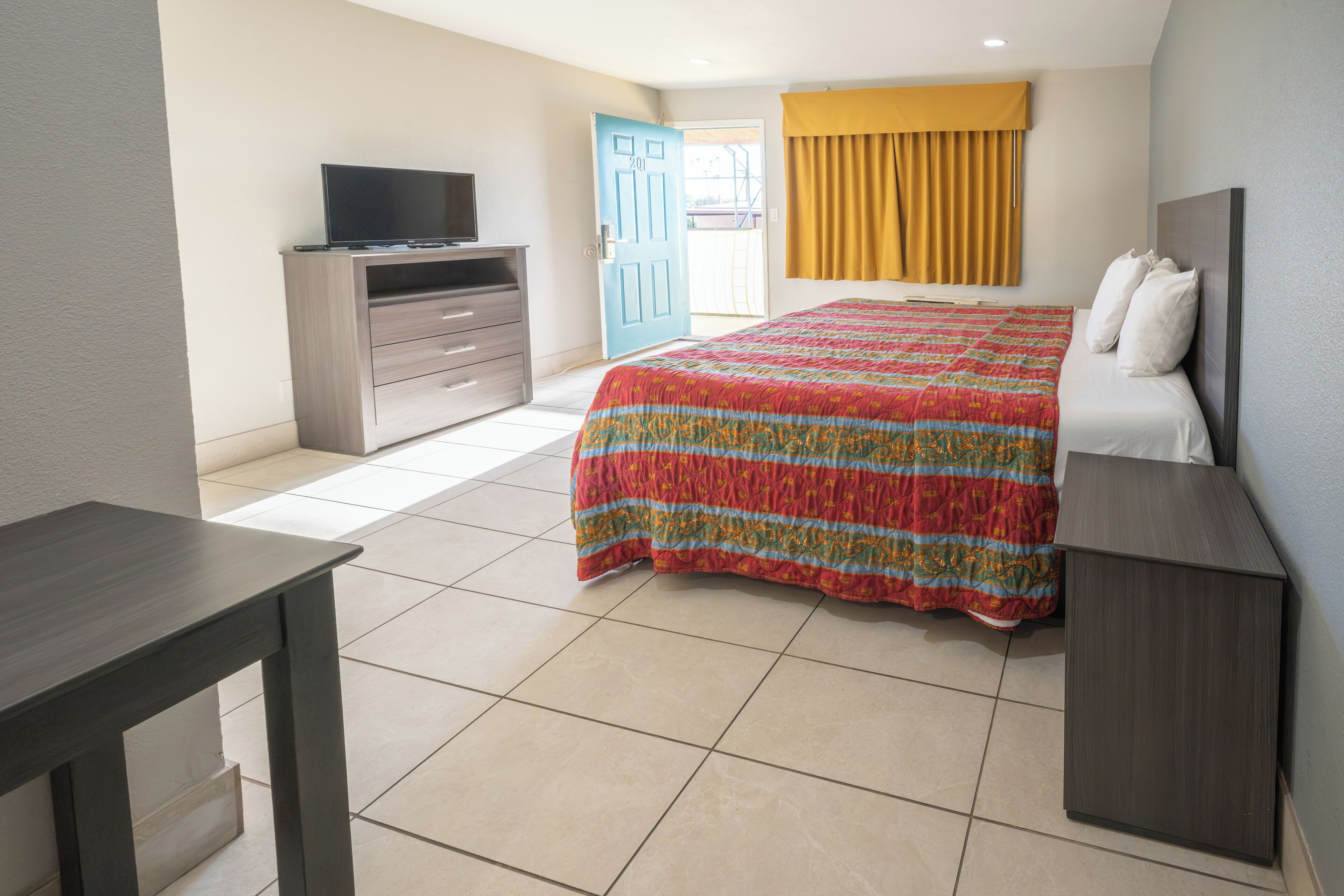 A hotel room with a bed, a TV on a dresser, a desk, and a door leading to an outdoor area.