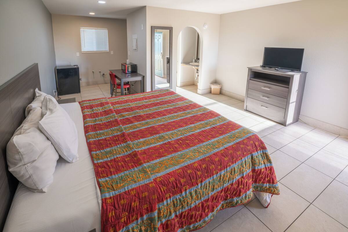 Habitación de hotel con una cama grande, un escritorio con silla, un televisor y un baño visible a través de un arco.