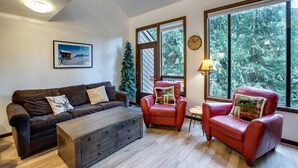 A living room with a brown leather sofa, a wooden coffee table, and two red leather armchairs. There is a large window with a view of trees and a clock on the wall. - Snowater Condo - You'll be Sitting in the Lap of Luxury (Deming)