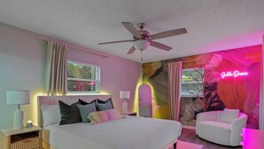 Ein Schlafzimmer mit einem Bett, Nachttischen, einem Deckenventilator, einem Fenster mit Jalousien, einem Neonlichtschild und einem Stuhl.