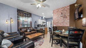 A living room with a sofa, coffee table, and dining table with chairs. There is a ceiling fan, a brick wall, and a kitchen in the background.