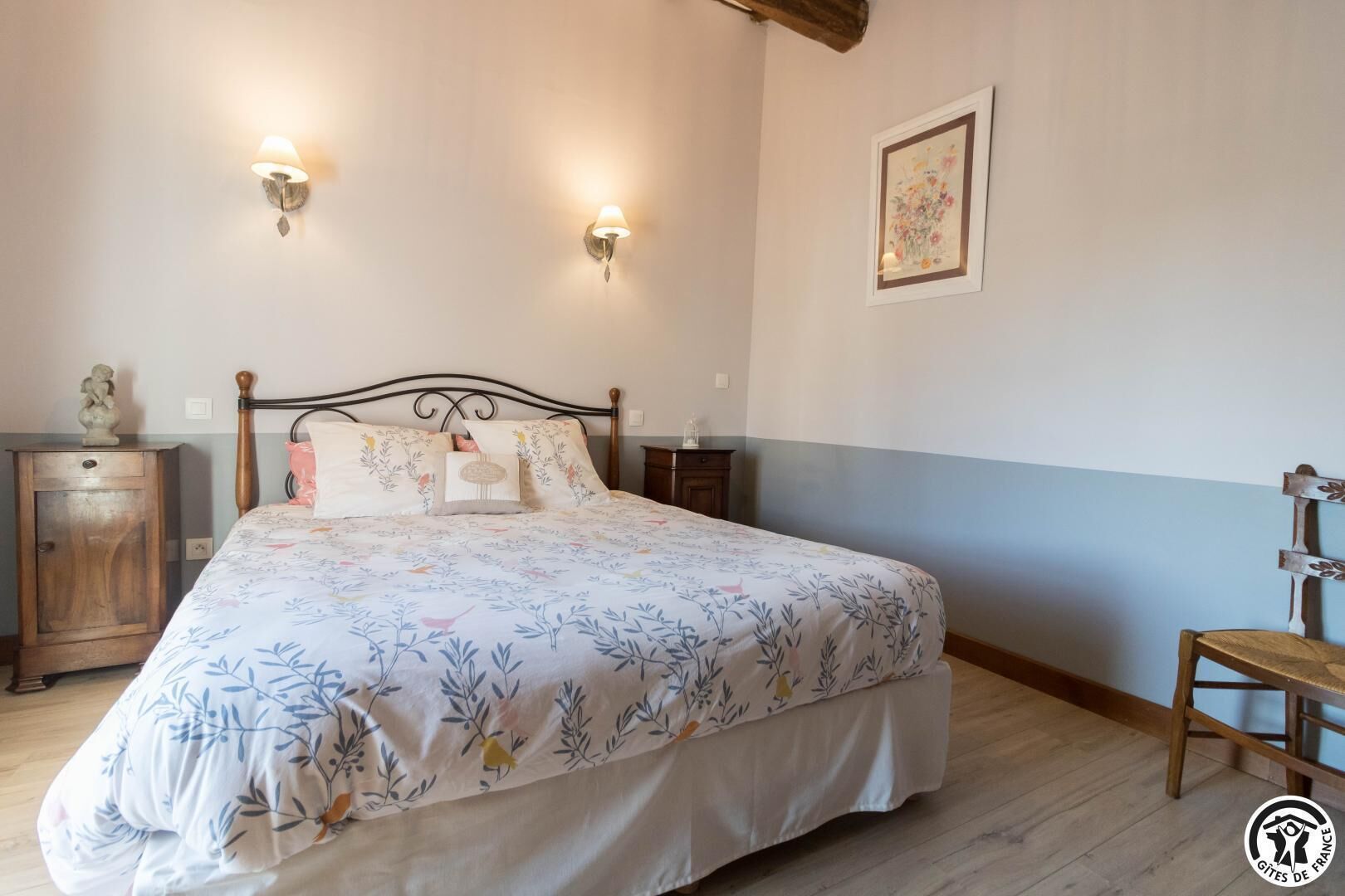 A bedroom with a bed, two bedside tables, a nightstand, a wooden chair, and a wall-mounted light fixture.
