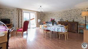 Un comedor con una mesa lista para cuatro personas, rodeada de sillas de madera. Hay un televisor montado en la pared, y una pared de piedra al fondo.