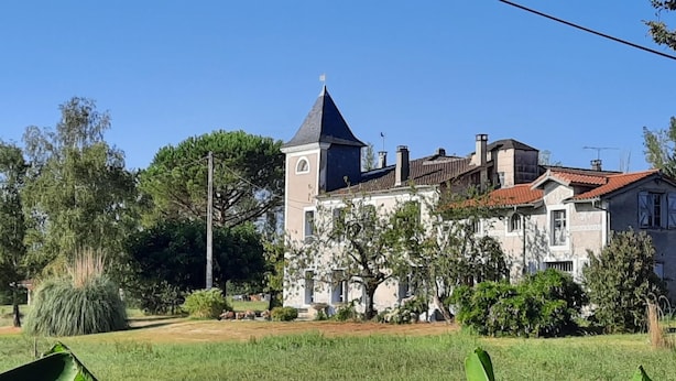 Ein großes, historisches Gebäude mit einem markanten Turm und zahlreichen Schornsteinen, umgeben von üppigem Grün und Bäumen.