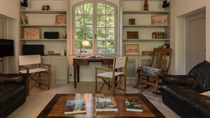 Un salon chaleureux avec un bureau en bois, deux fauteuils, une table basse avec des livres et une grande fenêtre donnant sur un paysage verdoyant.