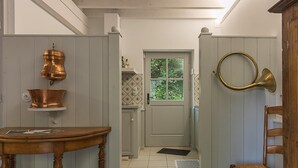 Un couloir étroit avec une table en bois, une bouilloire en laiton, une porte avec une fenêtre et une chaise en bois.