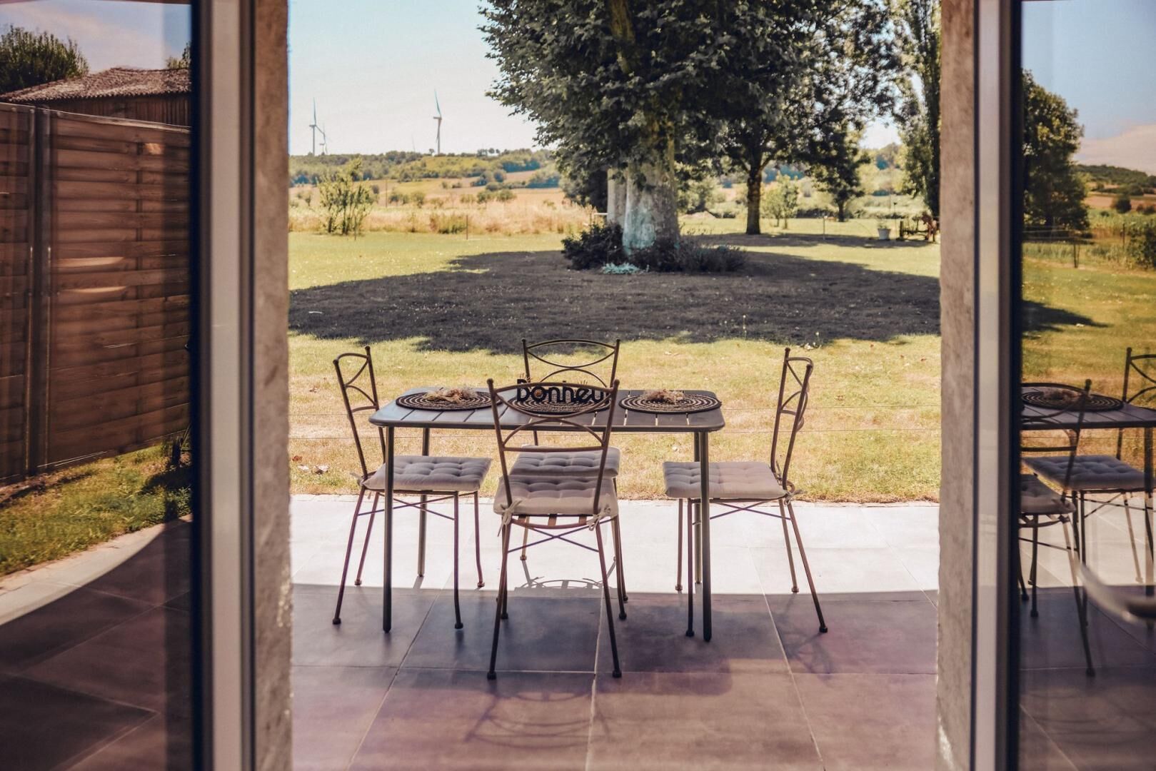 An outdoor patio with a round table and four chairs, a wooden fence, and a view of a grassy field with trees and wind turbines in the distance.