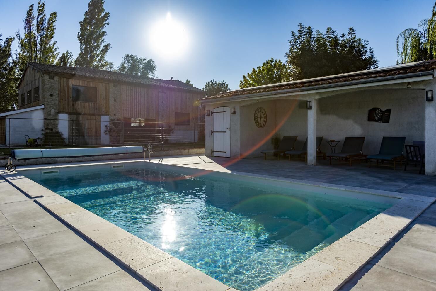 En rektangulær swimmingpool med klart vand, omgivet af en flisbelagt terrasse. Der er liggestole og et overdækket siddeområde med et ur på væggen.