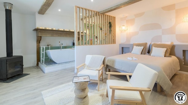 A modern bedroom with a large bed, two chairs, a wooden floor, a fireplace, and a built-in shelf with plants.