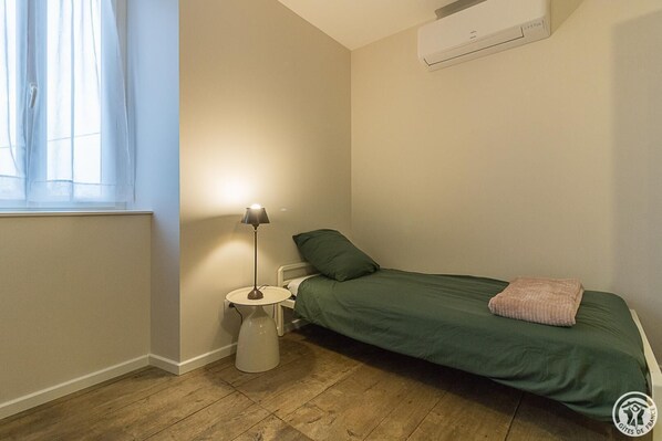A small bedroom with a single bed, a bedside table with a lamp, a radiator, and an air conditioning unit.