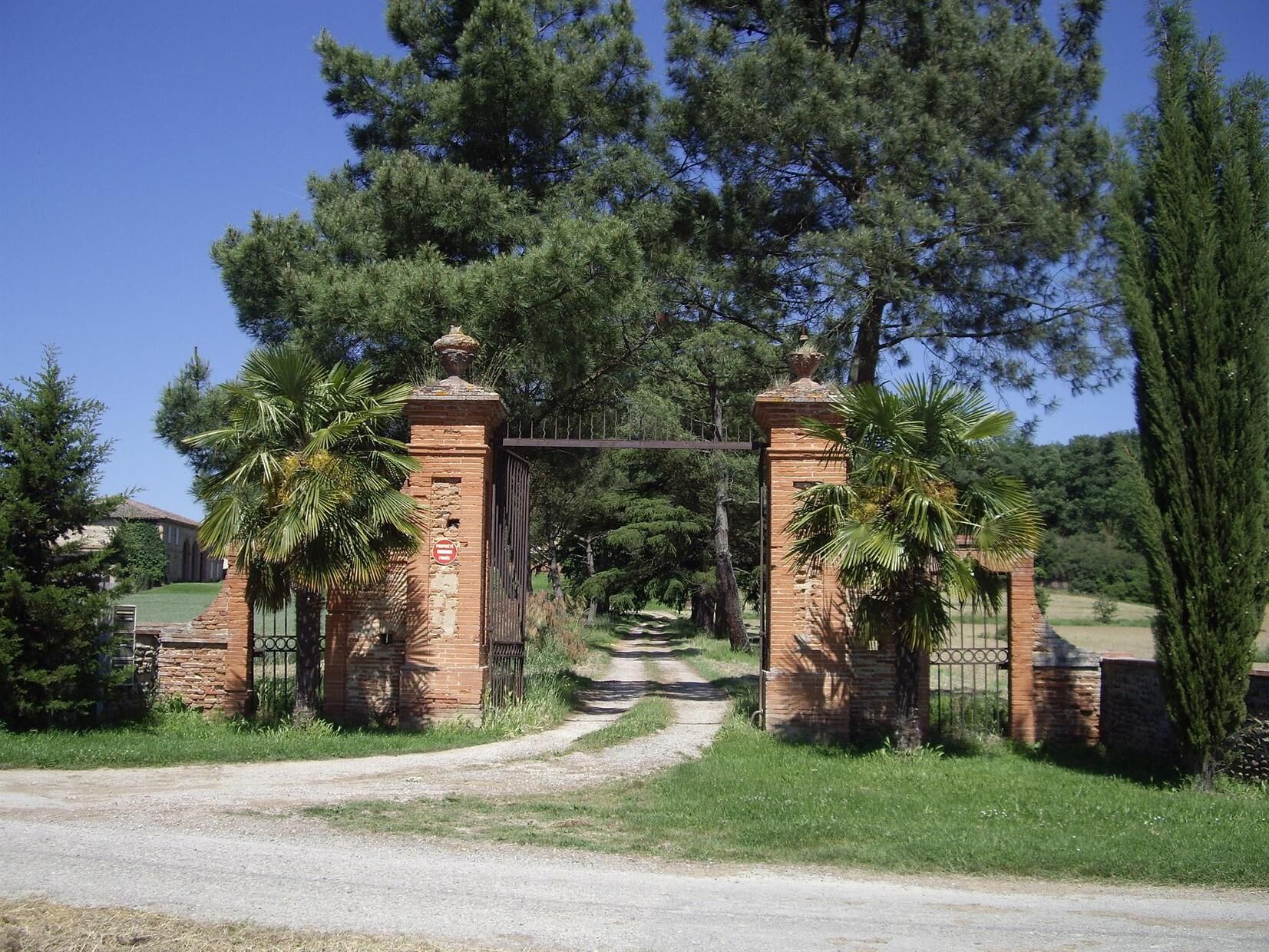 A rustic gate with brick pillars and a metal gate, leading to a path surrounded by lush greenery and tall trees.