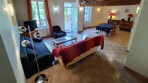 A spacious living area with a sofa, a coffee table, and a dining table set for four. There is a television and a lamp in the room.